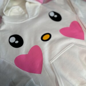 Puede incluir: Sudadera blanca con un lazo rosa, ojos negros, una nariz amarilla y dos corazones rosas en la parte delantera.