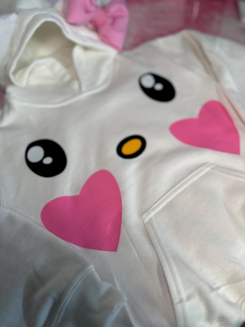 Puede incluir: Sudadera blanca con un lazo rosa, ojos negros, una nariz amarilla y dos corazones rosas en la parte delantera.