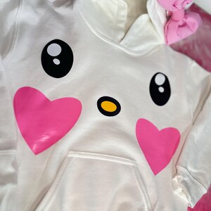Puede incluir: Sudadera blanca con un dise&ntilde;o de cara de dibujos animados con corazones rosas, ojos negros y una nariz amarilla. La sudadera tiene un lazo rosa en la capucha.