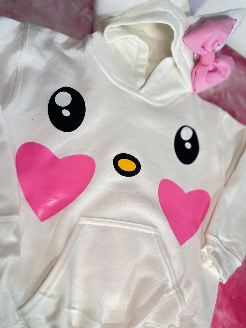 Puede incluir: Sudadera blanca con un dise&ntilde;o de cara de dibujos animados con corazones rosas, ojos negros y una nariz amarilla. La sudadera tiene un lazo rosa en la capucha.