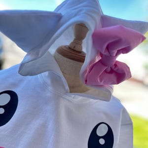 Puede incluir: Sudadera con capucha blanca con una cara de dibujos animados y un lazo rosa. La sudadera tiene ojos negros grandes, una nariz amarilla y una boca rosa.