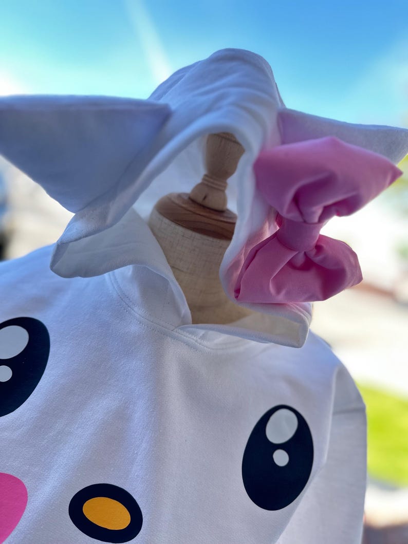 Puede incluir: Sudadera con capucha blanca con una cara de dibujos animados y un lazo rosa. La sudadera tiene ojos negros grandes, una nariz amarilla y una boca rosa.