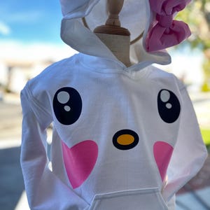 Puede incluir: Sudadera con capucha blanca con un dise&ntilde;o de cara de gato de dibujos animados. El gato tiene mejillas rosadas, ojos negros, una nariz amarilla y un lazo rosa en la capucha.