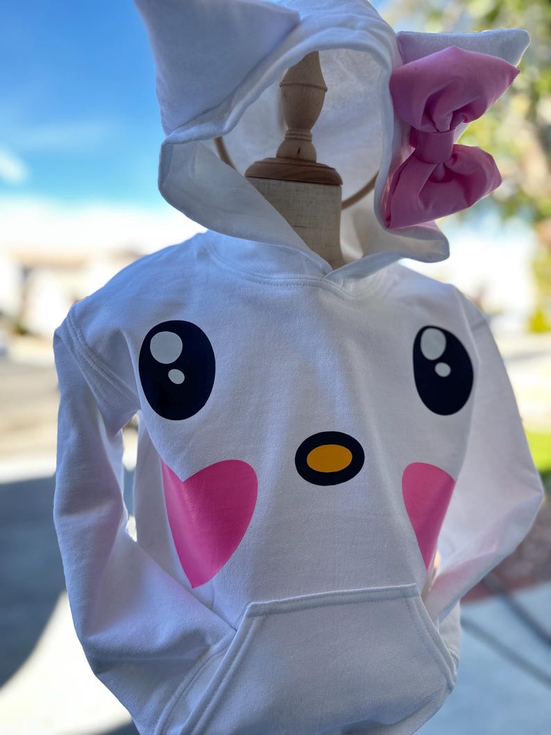 Puede incluir: Sudadera con capucha blanca con un dise&ntilde;o de cara de gato de dibujos animados. El gato tiene mejillas rosadas, ojos negros, una nariz amarilla y un lazo rosa en la capucha.