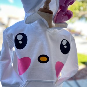 Puede incluir: Sudadera con capucha blanca con un dise&ntilde;o de cara de animal de dibujos animados. La cara tiene ojos negros grandes, una nariz amarilla y mejillas rosadas. La capucha tiene orejas blancas y un lazo rosa.