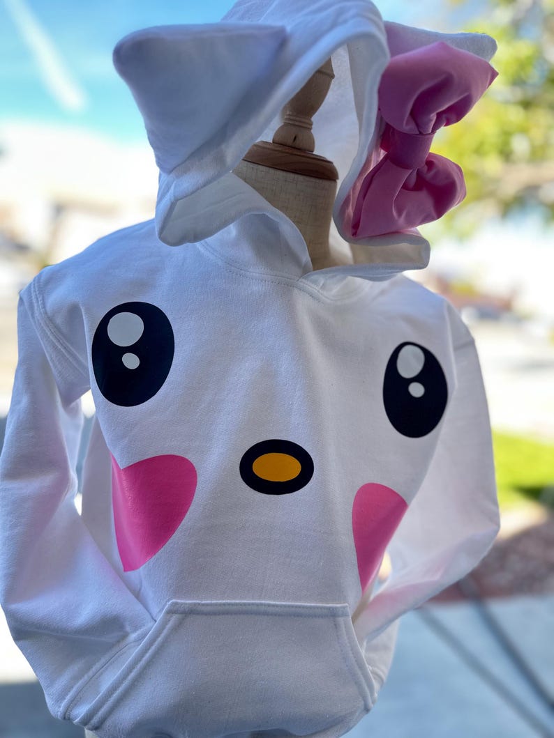 Puede incluir: Sudadera con capucha blanca con un dise&ntilde;o de cara de animal de dibujos animados. La cara tiene ojos negros grandes, una nariz amarilla y mejillas rosadas. La capucha tiene orejas blancas y un lazo rosa.