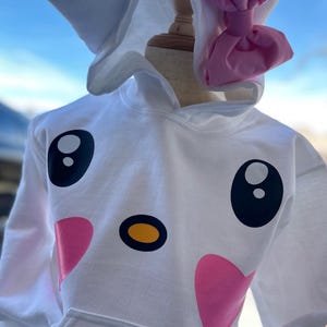 Puede incluir: Sudadera con capucha blanca con un dise&ntilde;o de cara de dibujos animados con ojos negros, nariz amarilla y corazones rosas. La capucha tiene orejas de gato blancas y un lazo rosa.
