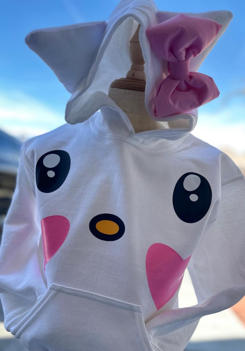 Puede incluir: Sudadera con capucha blanca con un dise&ntilde;o de cara de dibujos animados con ojos negros, nariz amarilla y corazones rosas. La capucha tiene orejas de gato blancas y un lazo rosa.