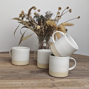 May include: Four white ceramic mugs with a speckled finish and a beige base are arranged on a wooden surface. A glass jar with dried flowers is in the background. The mugs have a simple, modern design.