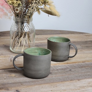 May include: Two grey ceramic mugs with a green glaze inside. The mugs are sitting on a wooden table.