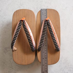 May include: Wooden geta sandals with light brown platforms and pink and black checkered straps. A ruler shows the sandals are approximately 9.8 inches long. The traditional Japanese footwear is displayed on a white surface.