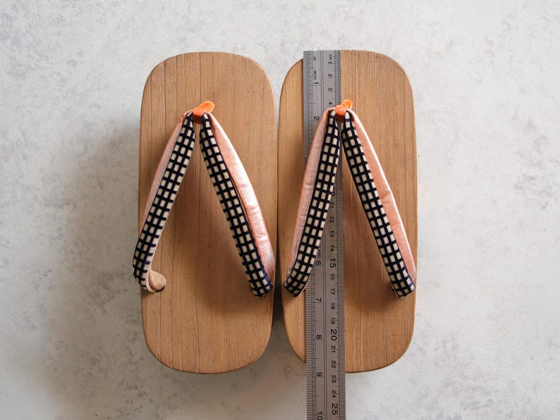 May include: Wooden geta sandals with light brown platforms and pink and black checkered straps. A ruler shows the sandals are approximately 9.8 inches long. The traditional Japanese footwear is displayed on a white surface.