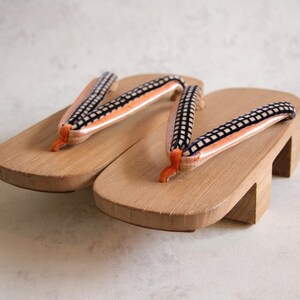 May include: A pair of traditional Japanese geta sandals. The wooden platforms are light brown, with elevated heels. The straps are black and white checkered, with orange and white accents. The sandals are designed for outdoor wear.