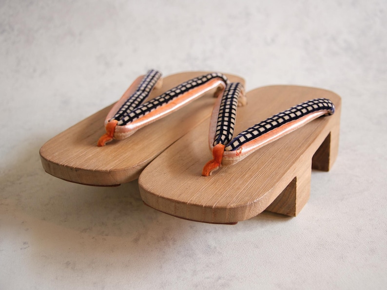 May include: A pair of traditional Japanese geta sandals. The wooden platforms are light brown, with elevated heels. The straps are black and white checkered, with orange and white accents. The sandals are designed for outdoor wear.