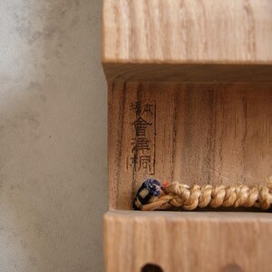 May include: Close-up of a wooden object with Japanese characters carved into the wood. A braided cord with blue and black accents is visible. The wood has a natural, light brown color.