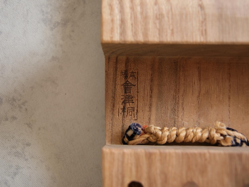 May include: Close-up of a wooden object with Japanese characters carved into the wood. A braided cord with blue and black accents is visible. The wood has a natural, light brown color.