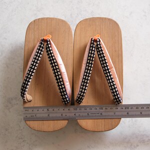 May include: Pair of wooden geta sandals with light pink straps and black and white checkered accents. The sandals are approximately 10 inches long, with a ruler placed across them for measurement. The geta are a traditional Japanese footwear.