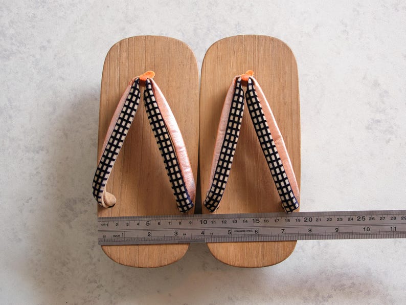 May include: Pair of wooden geta sandals with light pink straps and black and white checkered accents. The sandals are approximately 10 inches long, with a ruler placed across them for measurement. The geta are a traditional Japanese footwear.