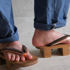May include: Wooden geta sandals with black and white straps and orange accents. The sandals are worn with blue denim jeans. The geta are elevated on two wooden blocks.