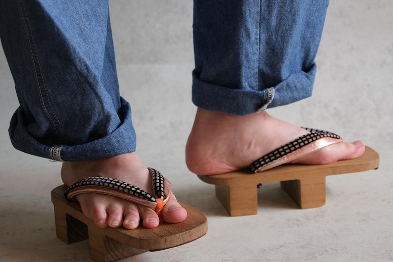 May include: Wooden geta sandals with black and white straps and orange accents. The sandals are worn with blue denim jeans. The geta are elevated on two wooden blocks.