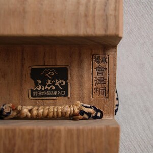 May include: Close-up of a wooden box with Japanese text and a woven cord. The box has a dark rectangular label with Japanese characters and a smaller rectangular label with vertical text. The cord is beige and blue.