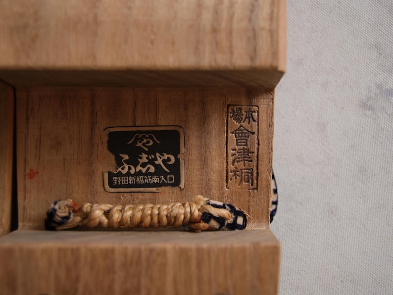 May include: Close-up of a wooden box with Japanese text and a woven cord. The box has a dark rectangular label with Japanese characters and a smaller rectangular label with vertical text. The cord is beige and blue.