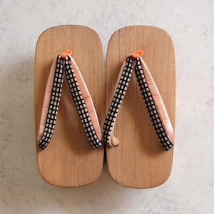 May include: A pair of traditional Japanese geta sandals. The wooden platforms are light brown, with peach-colored straps featuring a black and white checkered pattern. The straps are secured with small orange pegs.