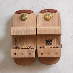 May include: Pair of traditional Japanese geta sandals made of light-colored wood. Each sandal features a raised platform, a woven thong, and decorative metal accents. The sandals have a natural wood grain finish.