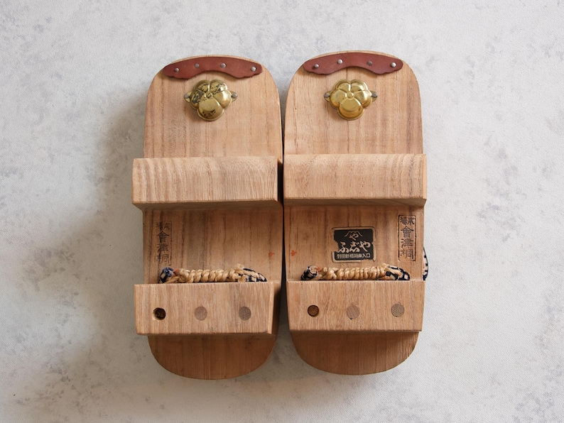 May include: Pair of traditional Japanese geta sandals made of light-colored wood. Each sandal features a raised platform, a woven thong, and decorative metal accents. The sandals have a natural wood grain finish.