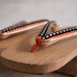 May include: Close-up of a pair of traditional Japanese geta sandals. The wooden soles are light brown, with orange and black patterned straps. The sandals are made of wood and fabric.