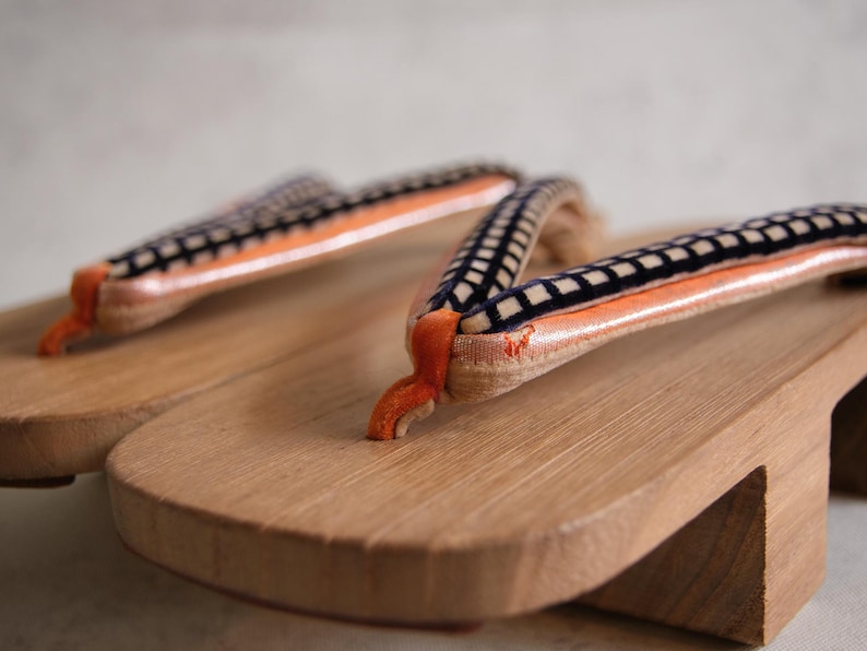 May include: Close-up of a pair of traditional Japanese geta sandals. The wooden soles are light brown, with orange and black patterned straps. The sandals are made of wood and fabric.