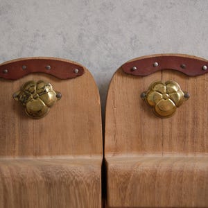 May include: Two wooden bookends with a light brown finish. Each bookend features a brass flower-shaped handle and a curved brown leather accent secured with small rivets. The bookends are designed to hold books upright.