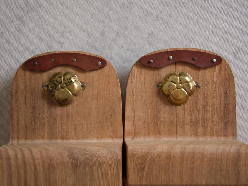 May include: Two wooden bookends with a light brown finish. Each bookend features a brass flower-shaped handle and a curved brown leather accent secured with small rivets. The bookends are designed to hold books upright.