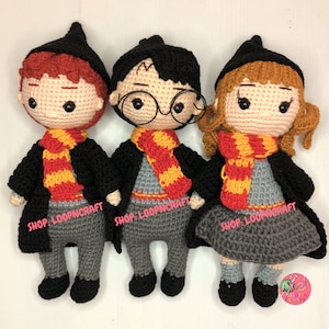 Puede incluir: Tres personajes de Harry Potter hechos a crochet, incluyendo Harry Potter, Ron Weasley y Hermione Granger. Cada personaje lleva una túnica negra, un suéter gris y una bufanda a rayas rojas y amarillas. Los personajes están de pie sobre un fondo blanco. SHOP: LOOPNCRAFT