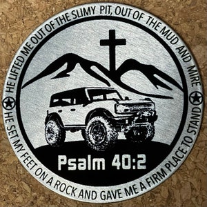 May include: A silver metal round coin with a black and white image of a jeep driving through a mountain pass. The coin has the text "HE LIFTED ME OUT OF THE SLIMY PIT, OUT OF THE MUD AND MIRE HE SET MY FEET ON A ROCK AND GAVE ME A FIRM PLACE TO STAND Psalm 40:2" around the edge.