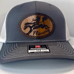 May include: Gray and white trucker hat with a brown leather patch featuring a horse and rider with the text "Flanery" in a brown oval. The hat is adjustable and has a Richardson 112 label.