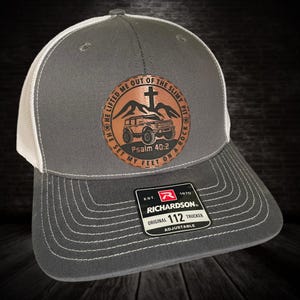 Peut inclure: Casquette de camionneur grise et blanche avec un patch en cuir marron représentant une jeep, des montagnes, une croix et le texte "HE LIFTED ME OUT OF THE SLIMY PIT HE SET MY FEET ON A ROCK Psalm 40:2". La casquette a une étiquette noire et rouge avec le texte "EST. 1970 RICHARDSON ORIGINAL 112 TRUCKER ADJUSTABLE".