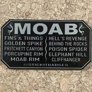 May include: Black sign with white text and graphics. The sign says "MOAB" and lists various trails in the Moab area, including "Fins & Things", "Hell's Revenge", "Golden Spike", "Behind the Rocks", "Pritchett Canyon", "Poison Spider", "Porcupine Rim", "Elephant Hill", "Moab Rim", and "Cliffhanger". The sign also includes the text "@STICKITBADGES".