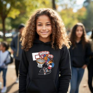 Puede incluir: Una sudadera con capucha negra con un gráfico de baloncesto con las palabras "HOOP LOOPS". El gráfico representa un personaje de dibujos animados jugando al baloncesto. La sudadera con capucha la lleva una persona con cabello castaño rizado.