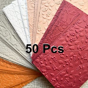 May include: A collection of 50 embossed paper sheets in various colors, including white, gray, orange, and shades of pink and red. The paper features floral and botanical patterns. The text "50 Pcs" is visible on the image.