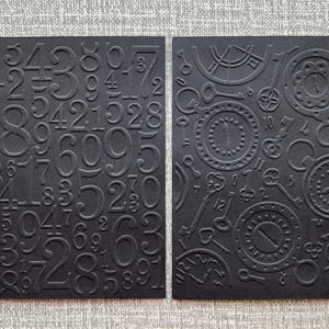 K&ouml;nnte beinhalten: Zwei schwarze Karten mit gepr&auml;gten Designs. Eine Karte zeigt erhabene Zahlen, die andere Zifferbl&auml;tter und Schl&uuml;ssel. Die Karten sind vor einem neutralen grauen Hintergrund platziert, was einen Kontrast erzeugt, der die komplizierten Details hervorhebt.