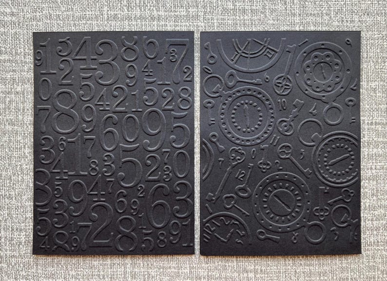 K&ouml;nnte beinhalten: Zwei schwarze Karten mit gepr&auml;gten Designs. Eine Karte zeigt erhabene Zahlen, die andere Zifferbl&auml;tter und Schl&uuml;ssel. Die Karten sind vor einem neutralen grauen Hintergrund platziert, was einen Kontrast erzeugt, der die komplizierten Details hervorhebt.