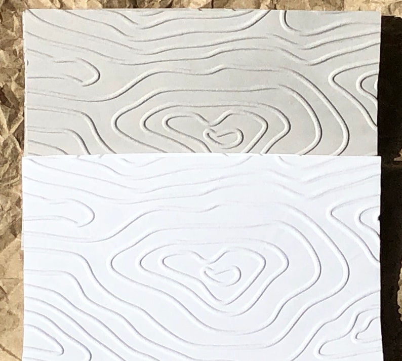 May include: Two textured paper samples, one light gray and one white, with a raised pattern of wavy lines resembling a topographic map.
