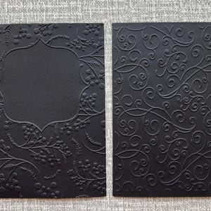 K&ouml;nnte beinhalten: Zwei schwarze Karten mit gepr&auml;gten Designs. Eine Karte zeigt ein Blumen- und Beerenmuster um einen leeren, dekorativen Rahmen. Die andere Karte hat ein wirbelndes, verziertes Design. Ideal f&uuml;r Einladungen oder Schreibwaren.