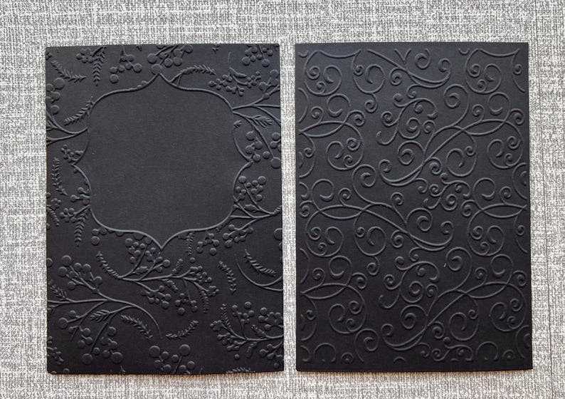 K&ouml;nnte beinhalten: Zwei schwarze Karten mit gepr&auml;gten Designs. Eine Karte zeigt ein Blumen- und Beerenmuster um einen leeren, dekorativen Rahmen. Die andere Karte hat ein wirbelndes, verziertes Design. Ideal f&uuml;r Einladungen oder Schreibwaren.