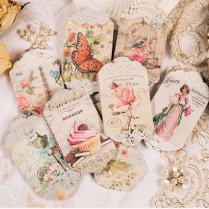 May include: Assortment of vintage-style decorative tags with lace trim. The tags feature images of a butterfly, a bird in a cage, a rose, a cupcake, a Parisian scene, and a woman in a dress. The tags are printed on a cream-colored background.