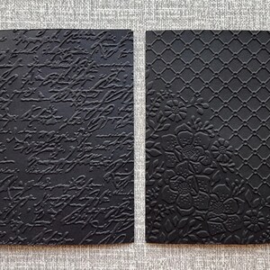 K&ouml;nnte beinhalten: Zwei schwarze rechteckige Karten mit gepr&auml;gten Designs. Eine zeigt Schrift, die andere ein Blumen- und Gittermuster. Die Karten sind wahrscheinlich f&uuml;r Bastelarbeiten, Schreibwaren oder Scrapbooking gedacht.