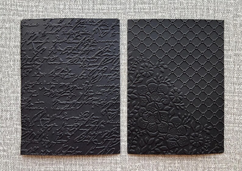 K&ouml;nnte beinhalten: Zwei schwarze rechteckige Karten mit gepr&auml;gten Designs. Eine zeigt Schrift, die andere ein Blumen- und Gittermuster. Die Karten sind wahrscheinlich f&uuml;r Bastelarbeiten, Schreibwaren oder Scrapbooking gedacht.