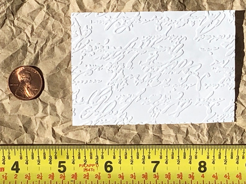 May include: A white textured sheet of paper with a repeating pattern of embossed swirls. The sheet is approximately 2 inches by 3 inches.