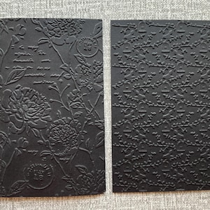 K&ouml;nnte beinhalten: Zwei schwarze, gepr&auml;gte Papierb&ouml;gen. Einer zeigt florale und Schriftmuster, der andere ein Muster aus Vogelk&auml;figen auf &Auml;sten. Das Papier hat ein strukturiertes Reliefdesign, ideal f&uuml;r Bastelarbeiten oder Schreibwaren.